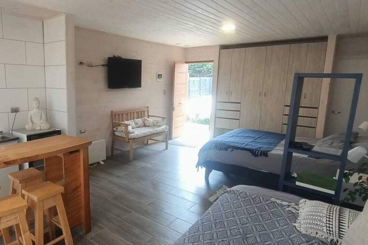 A compact, modern room with a kitchenette, a bed with a blue bedspread, a wooden bench, and a television mounted on the wall.