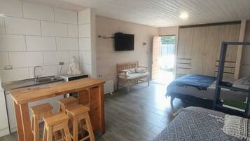A compact, modern room with a kitchenette, a bed with a blue bedspread, a wooden bench, and a television mounted on the wall.