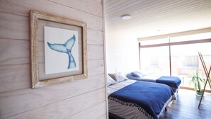 Habitación con paredes de madera, un cuadro de ballena enmarcado, dos camas con ropa de cama azul y un ventanal con vistas.