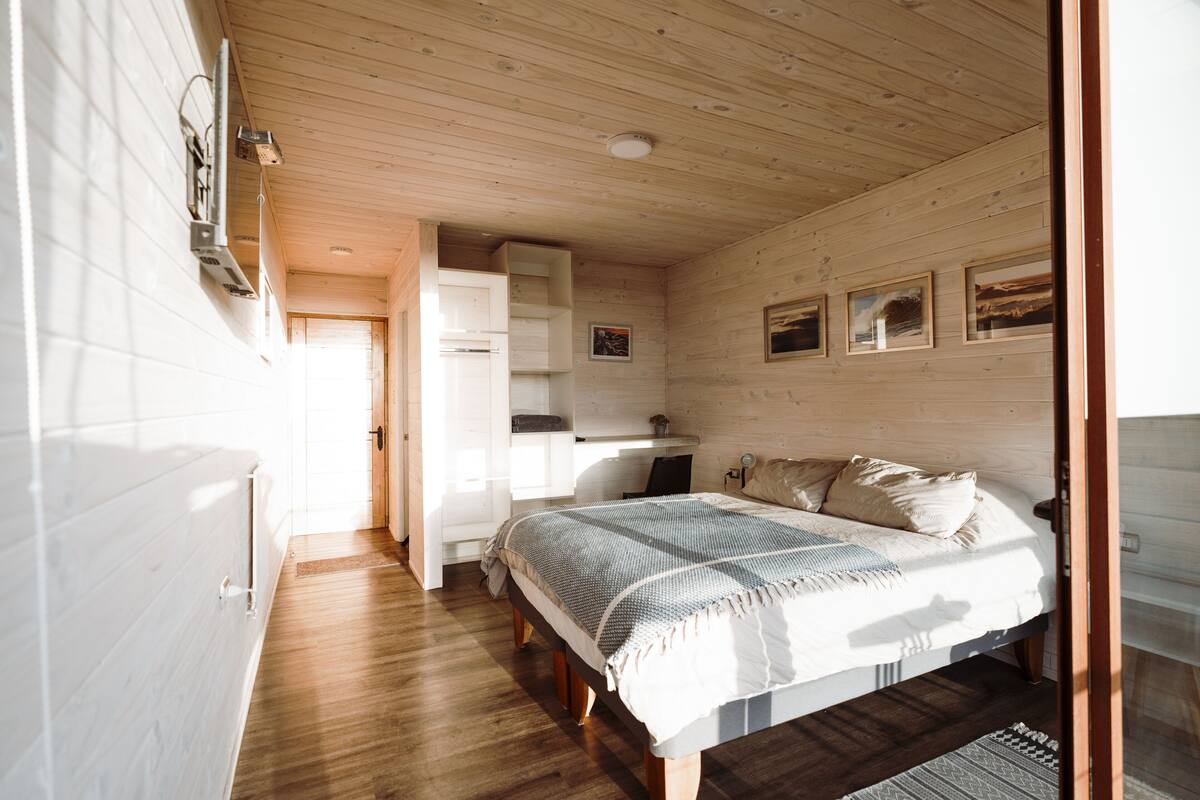 A bedroom with a wooden ceiling, a bed with a striped blanket, a desk with a chair, and a wall-mounted shelf.