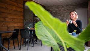 Una persona sentada a una mesa con una taza, en una habitación con paredes de madera y una pared de piedra al fondo.