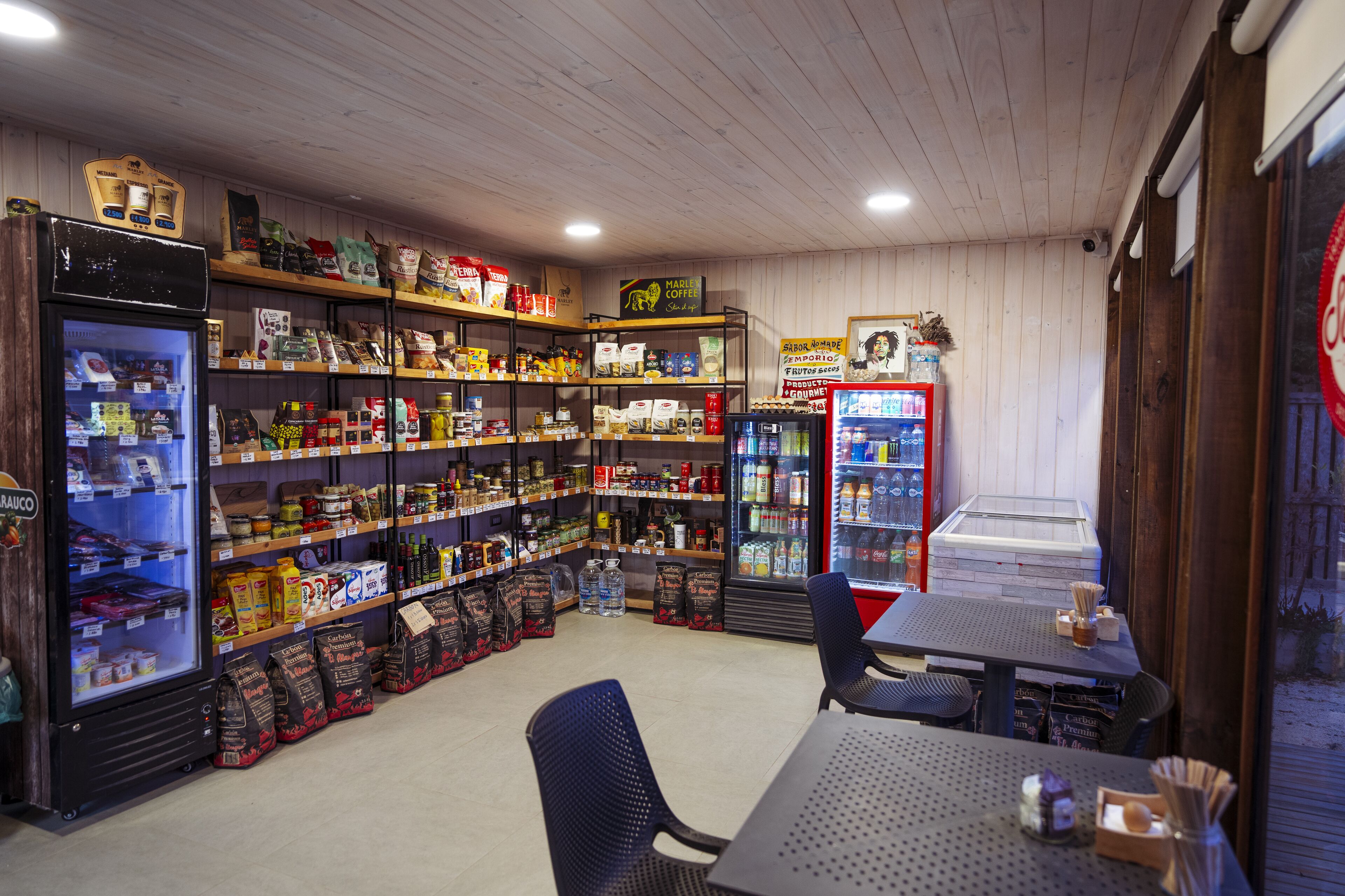 A store with shelves stocked with various products, a refrigerated display case, and a seating area with tables and chairs.