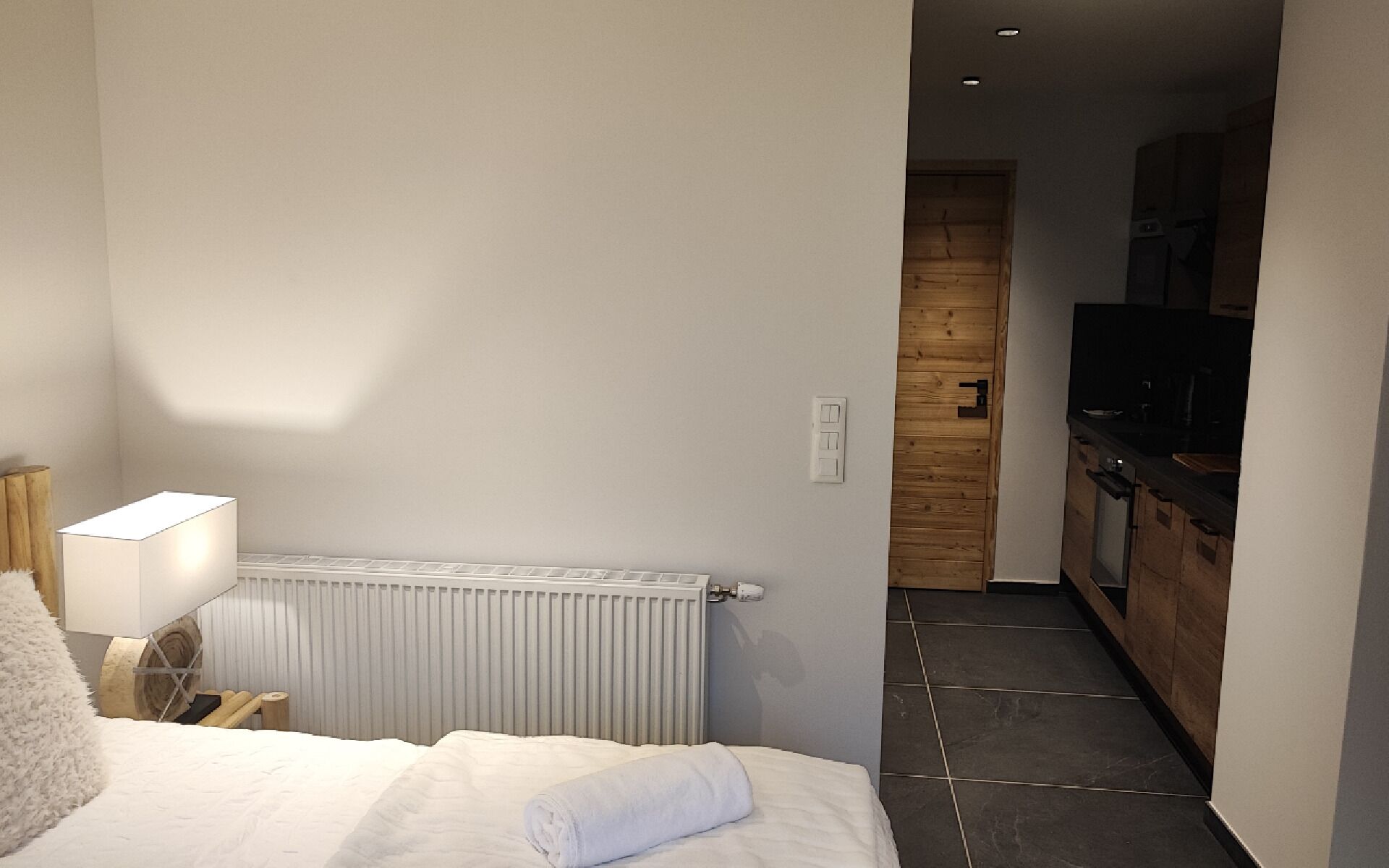 Ein Schlafzimmer mit einem Bett, einer Nachttischlampe und einer Heizung. Eine Holztür führt zu einem Küchenbereich mit einem Fernseher.