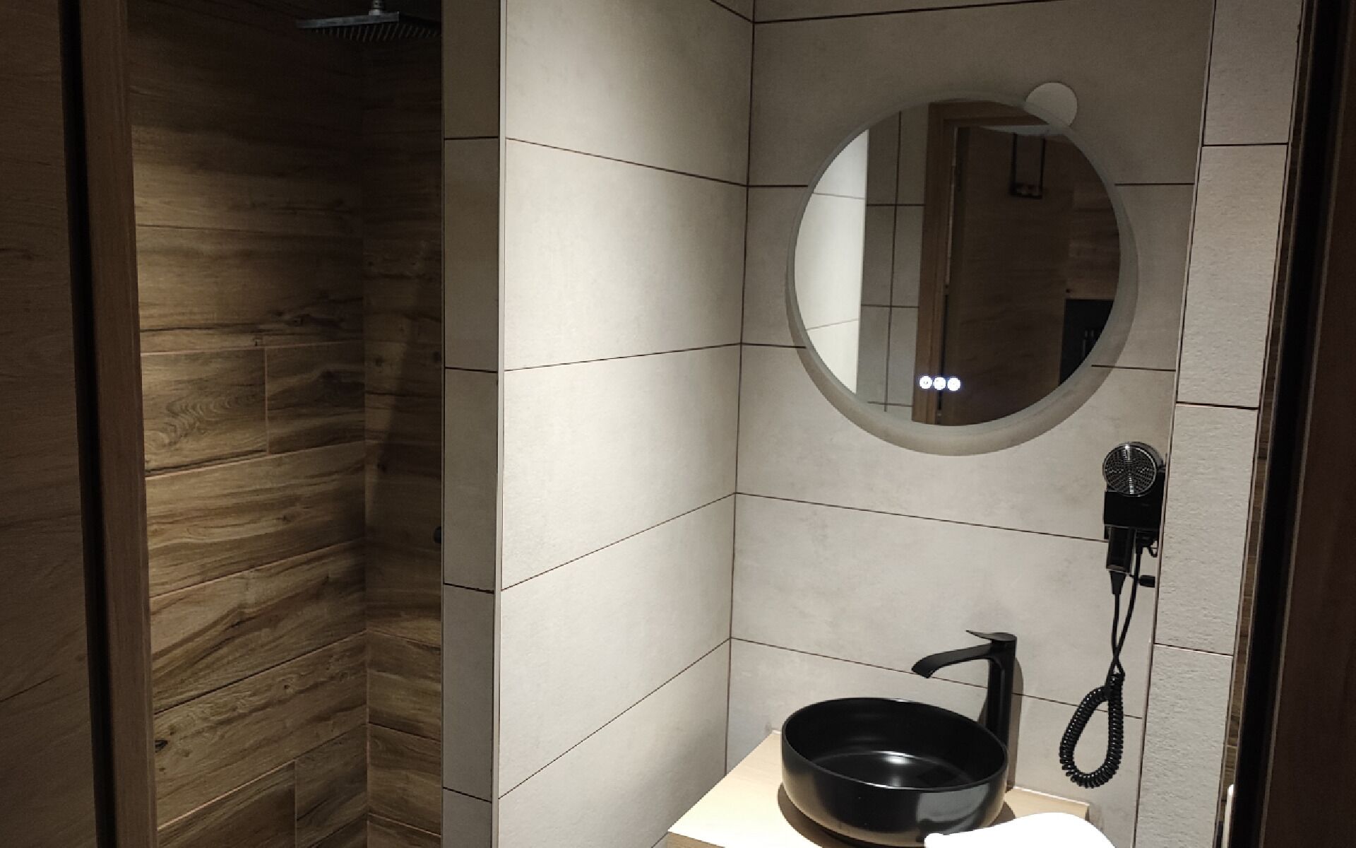 A modern bathroom with a wooden shower enclosure, a round mirror, and a black sink with a handheld showerhead.