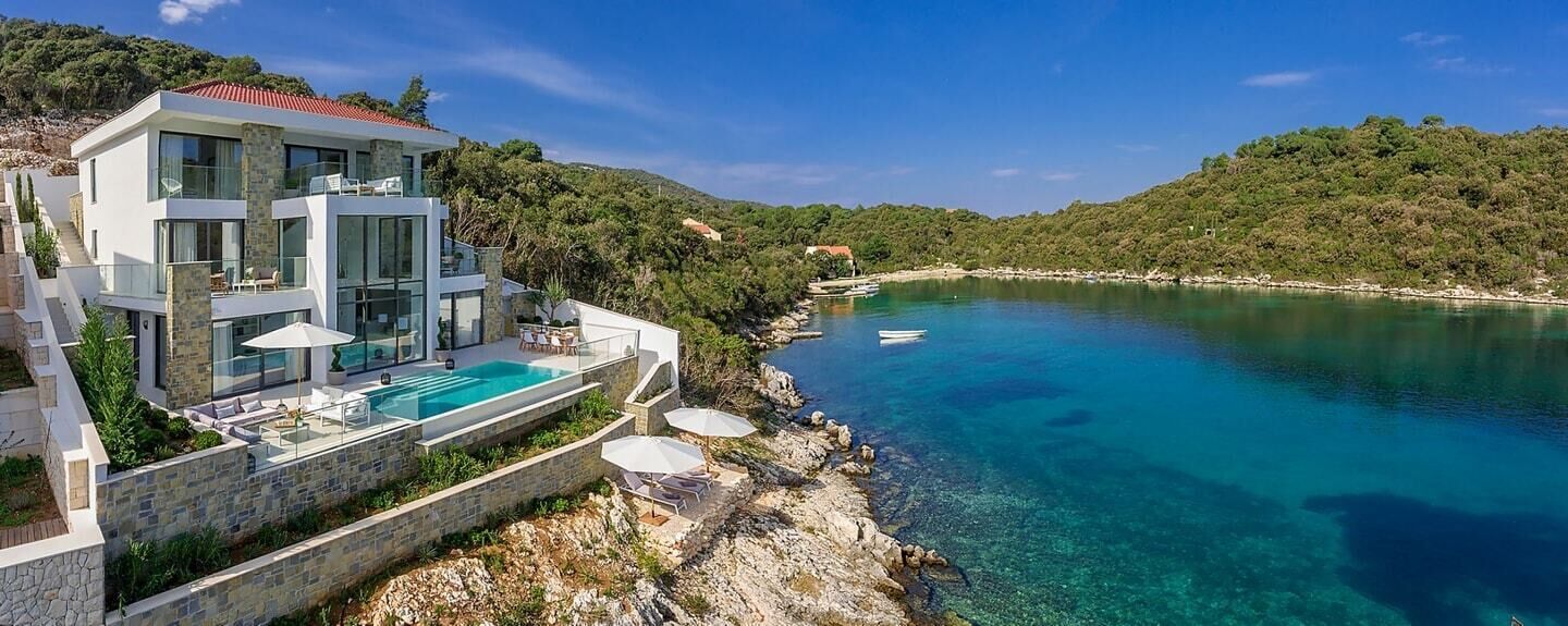 A modern villa with a pool, overlooking a clear blue lake and surrounded by hills.