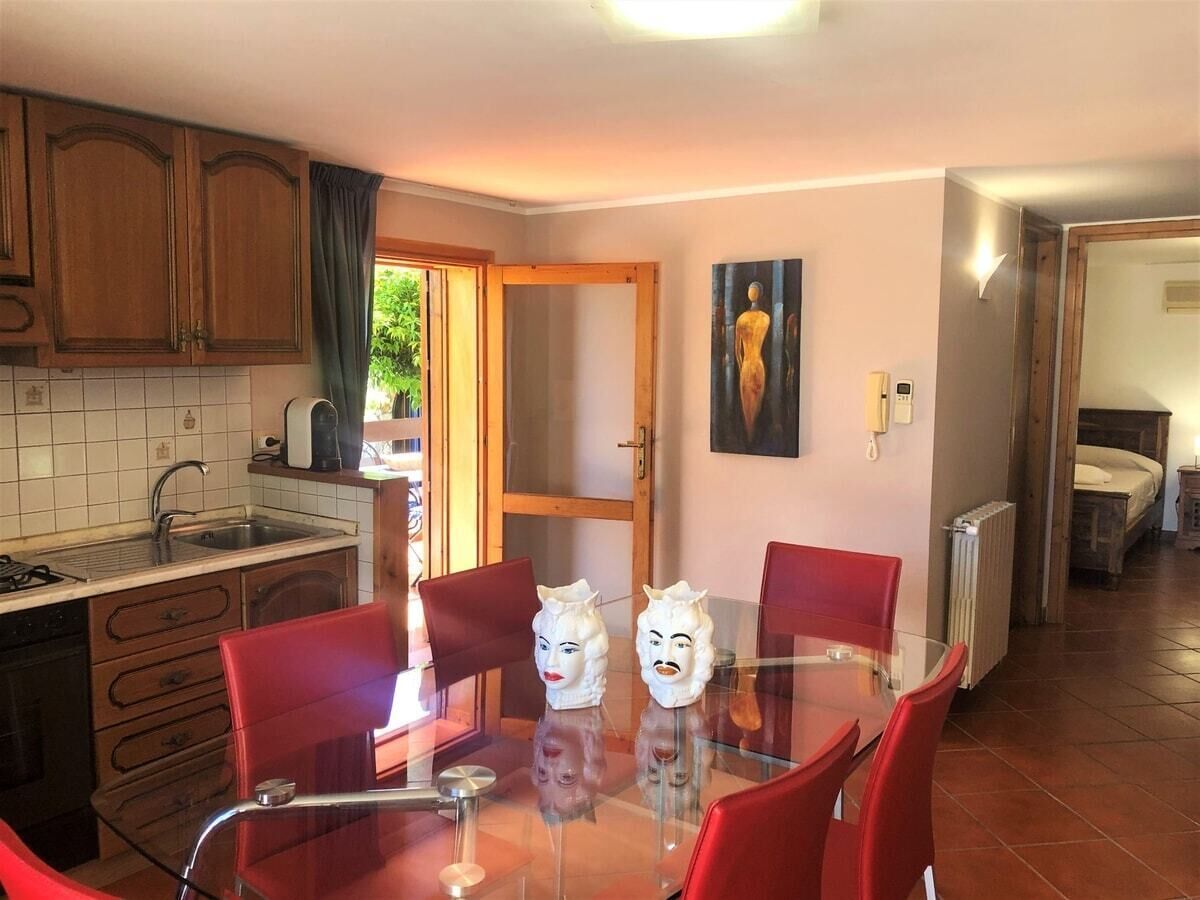 A dining area with a glass table and red chairs, a kitchen with wooden cabinets, and a painting on the wall.