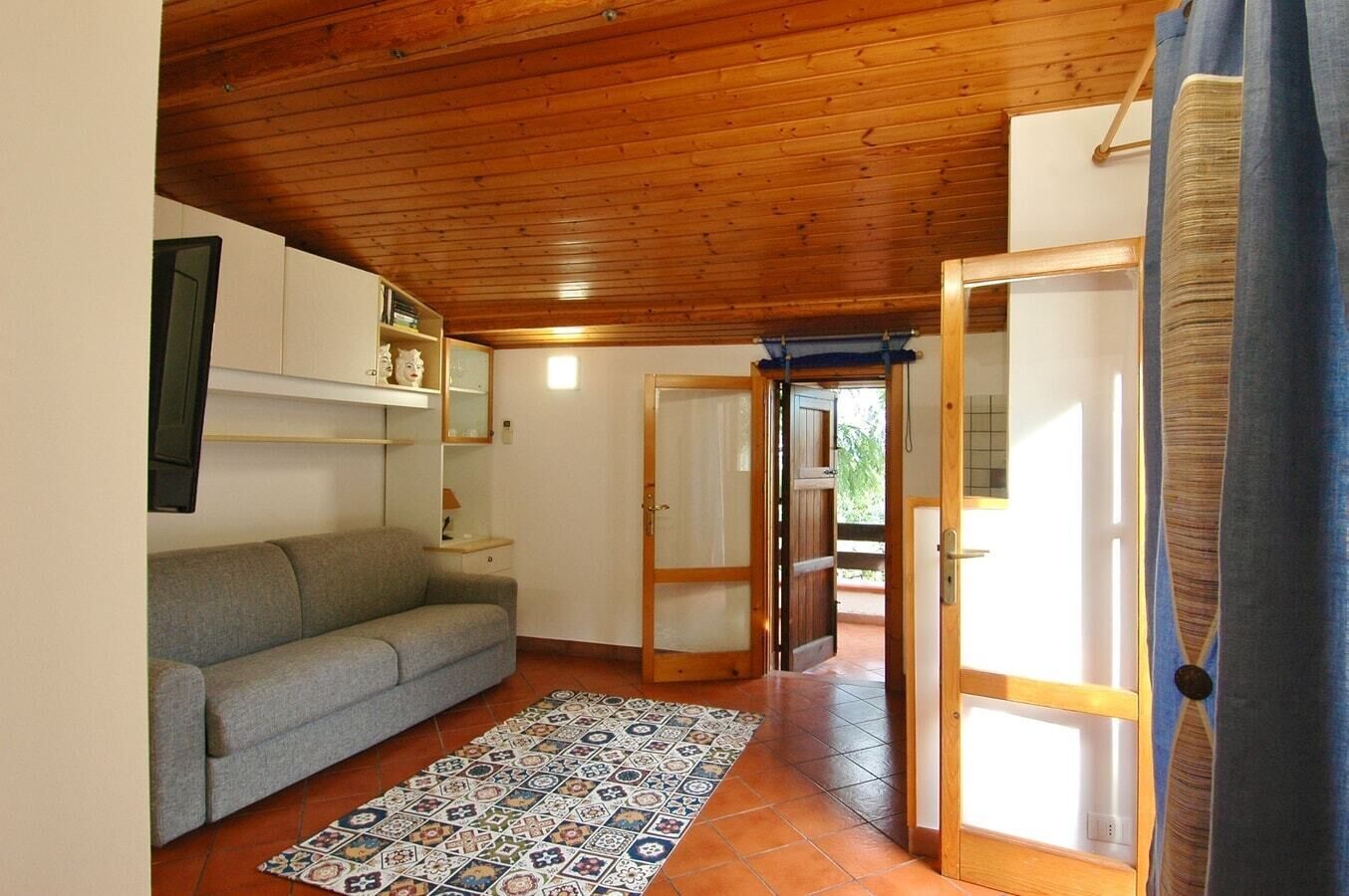 A cozy room with a grey sofa, a patterned rug, wooden ceiling, and a door leading outside.