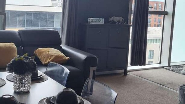 A modern living room with a dining table, black leather sofa, and a large window with curtains. - Immaculate 1-bed Apartment in Milton Keynes (Milton Keynes)