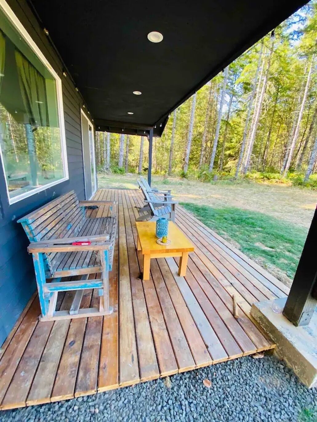 Une terrasse couverte, dotée de bancs en bois et d’une table, entourée d’une zone boisée.