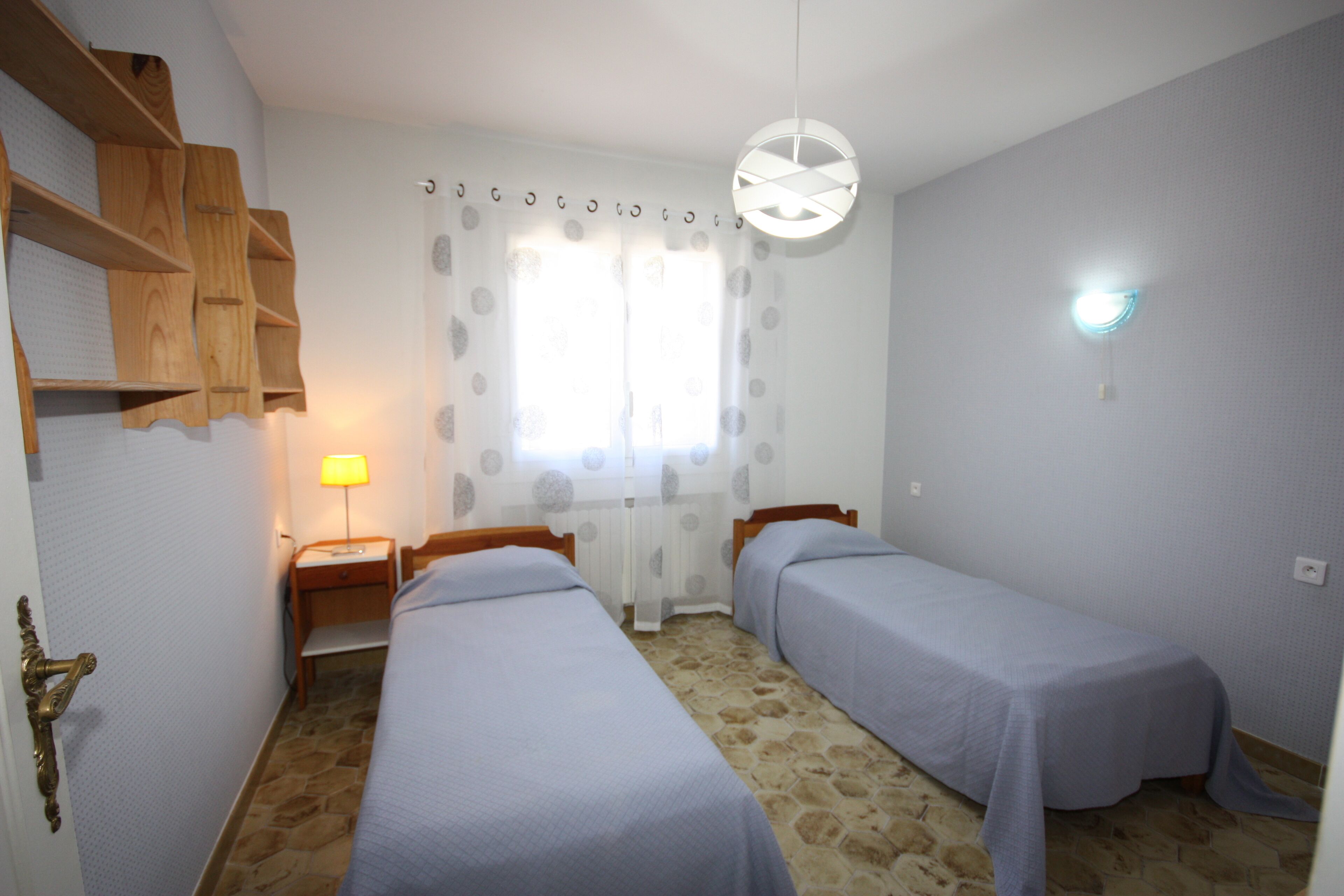 A room with two single beds, a wooden shelf unit, a nightstand with a lamp, and a window with a curtain.