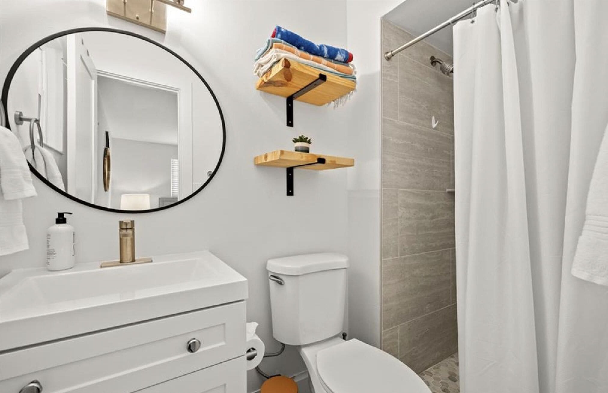 Une salle de bain avec un lavabo blanc, des toilettes, une douche avec un rideau blanc, un miroir rond et des étagères en bois où sont posées des serviettes et une petite plante.