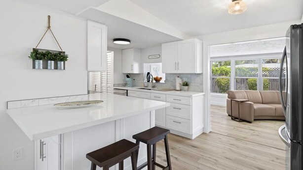 A modern kitchen with white cabinetry, a large island with stools, a hanging plant, and a view to a living area with a beige sofa.