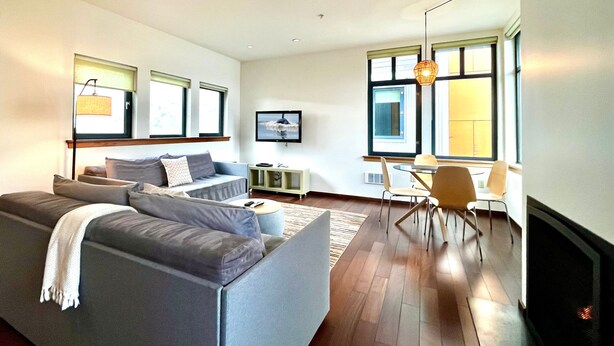 A modern living room with a grey sofa, a dining area with a table and chairs, and a fireplace. - ✨NEW✨ Bright in-town Penthouse with roof deck 😎 (Friday Harbor)