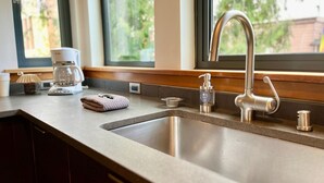 A kitchen with a stainless steel sink, a coffee maker, and a window with a view of greenery. - ✨NEW✨ Bright in-town Penthouse with roof deck 😎 (Friday Harbor)
