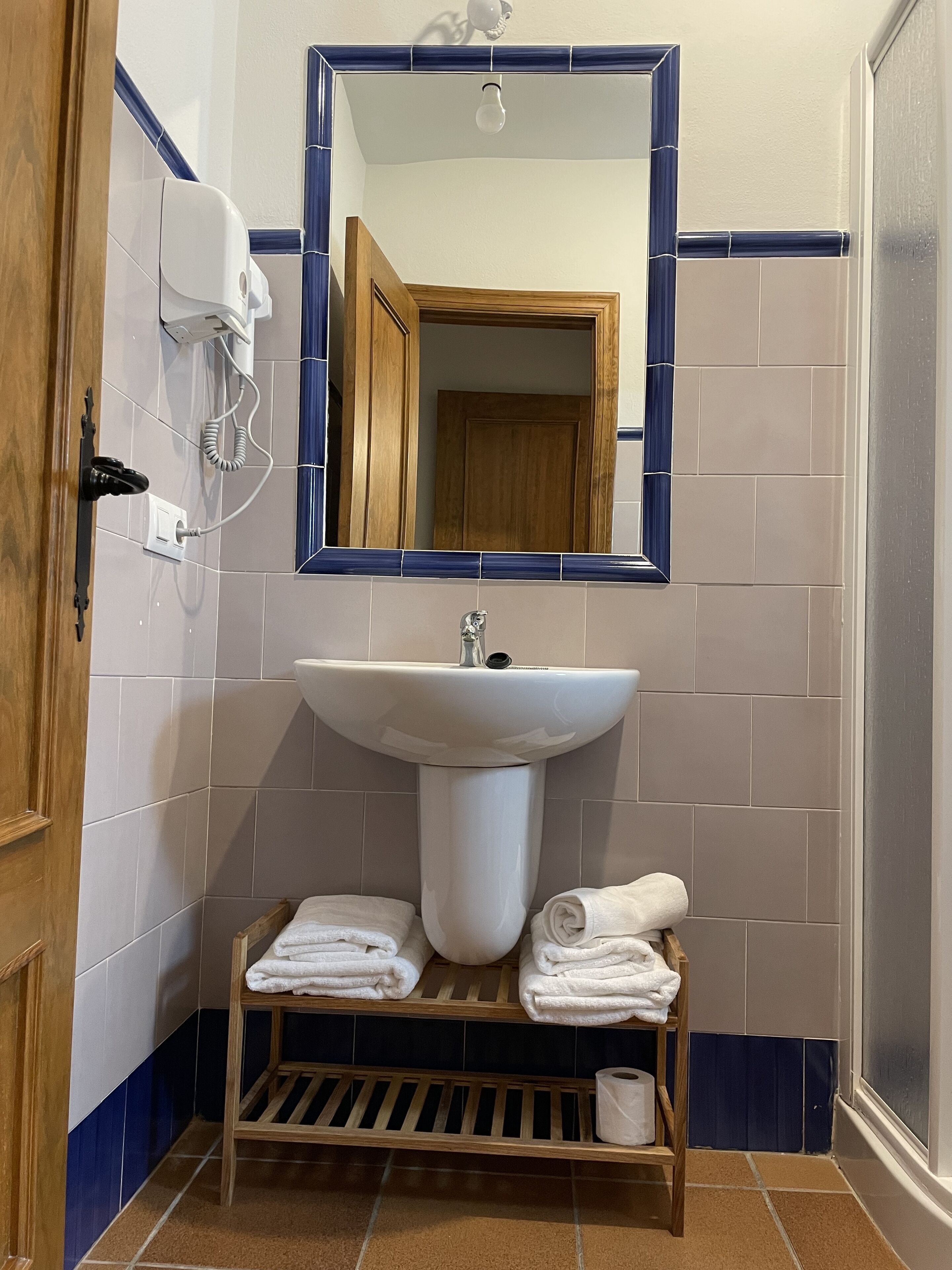 A bathroom with a white sink, a wooden shelf with towels, and a mirror with a blue frame.
