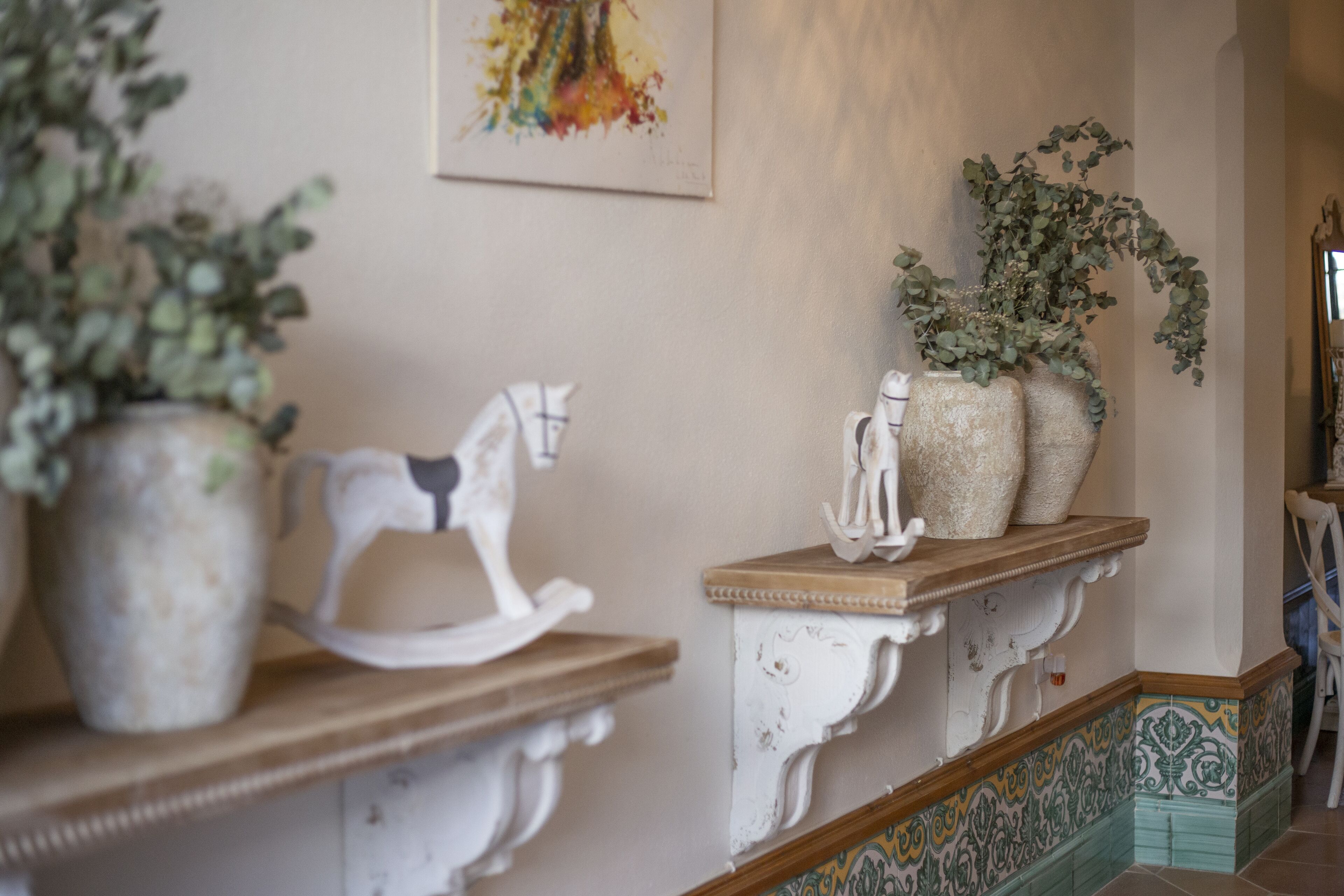 A hallway with a white rocking horse, two large vases with plants, and a colorful tiled floor.