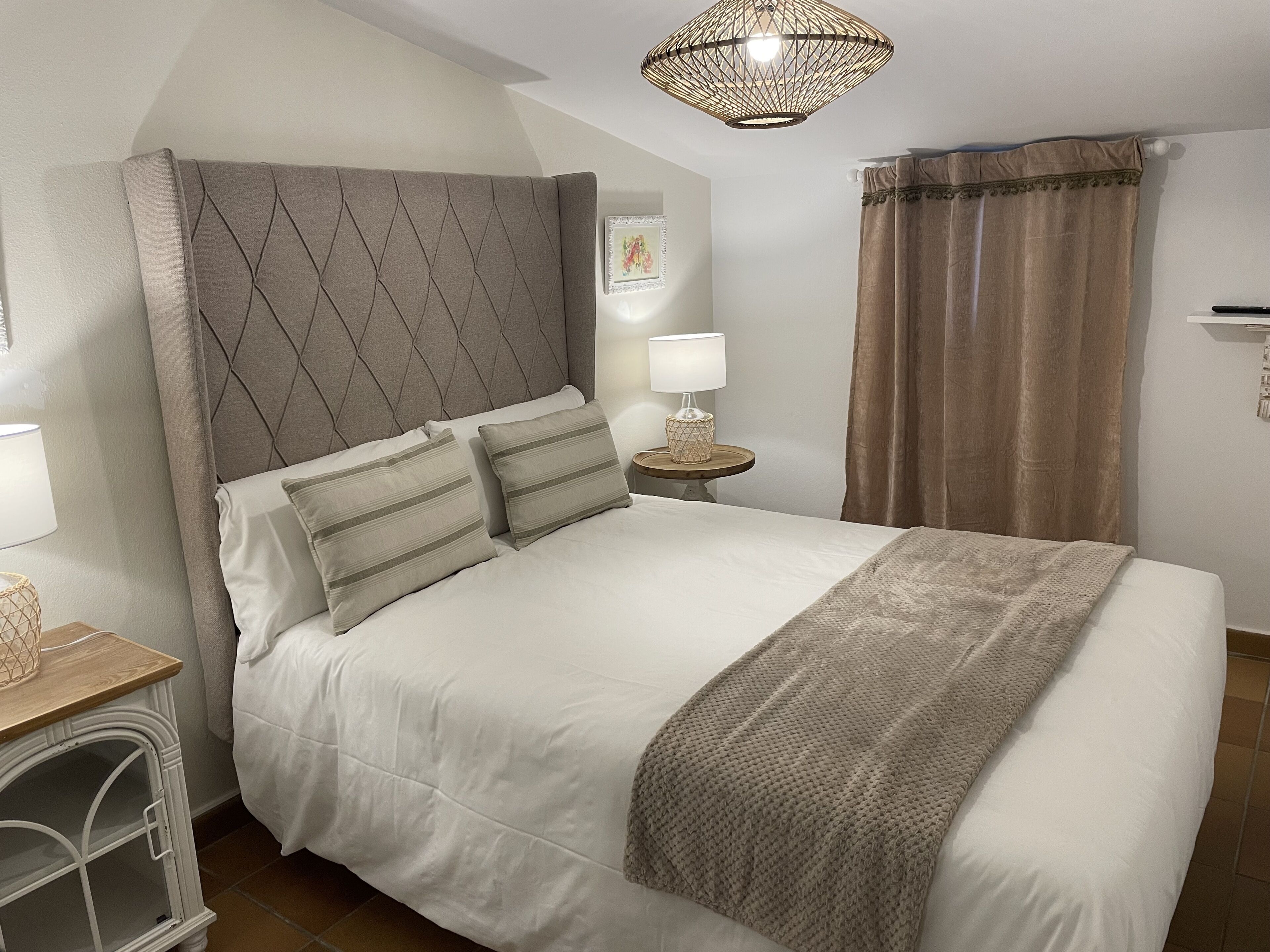 A neatly made bed with a grey headboard, a bedside table with a lamp, and a window with curtains.