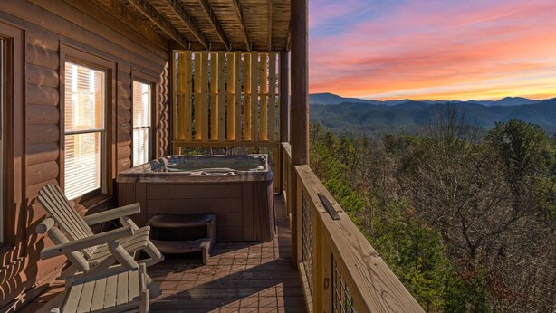 Een houten hut met een jacuzzi en terrasstoelen met uitzicht op een berglandschap tijdens de zonsondergang.