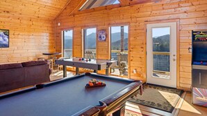 Een gezellige kamer met een pooltafel, een bank en een deur die leidt naar een buitenruimte met uitzicht op bergen.