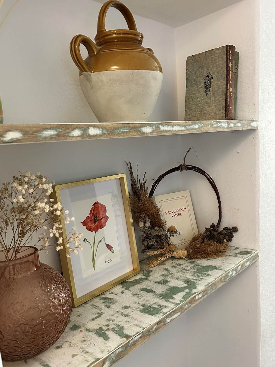 A shelf with a ceramic pot, a framed picture, a book, and a vase with dried flowers.