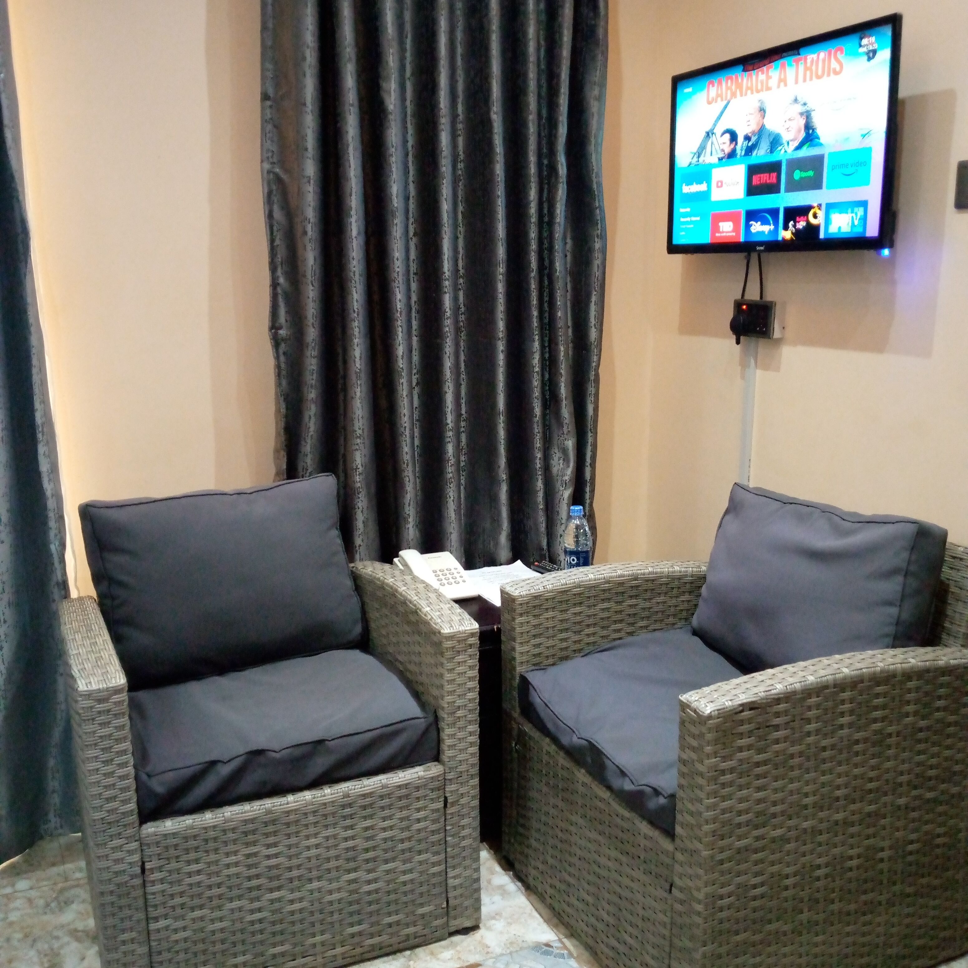 A room with two wicker chairs with dark cushions, a flat-screen TV mounted on the wall displaying a streaming service interface, and a curtain partially drawn to the side.