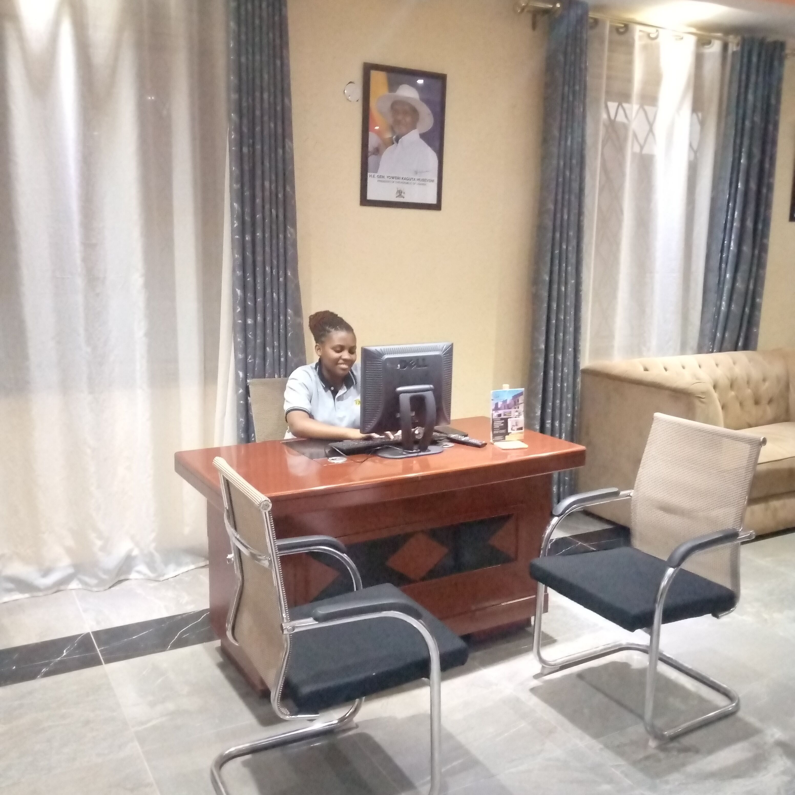 A person seated at a wooden desk with a computer, two chairs, and a framed picture on the wall.