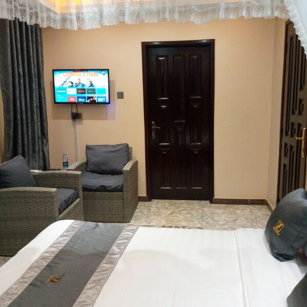 A hotel room with a bed, two wicker chairs, a TV displaying a streaming service interface, a dark wooden door, and a water bottle on the floor. - Hotel Zeptan (Jinja)