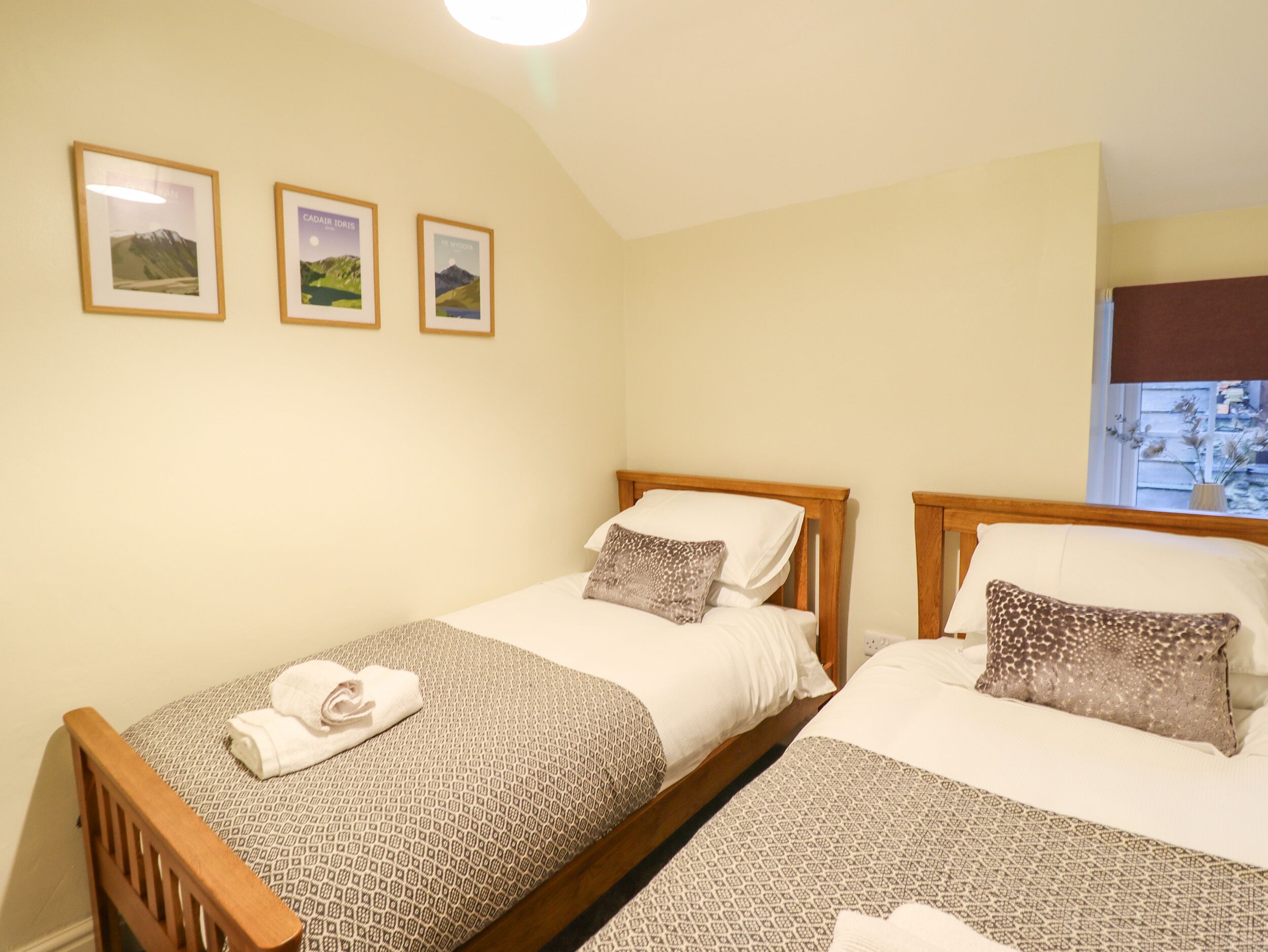 A bedroom with two single beds, each with patterned bedding and pillows, a wooden headboard, and a small bedside table. There are three framed pictures on the wall above the beds.