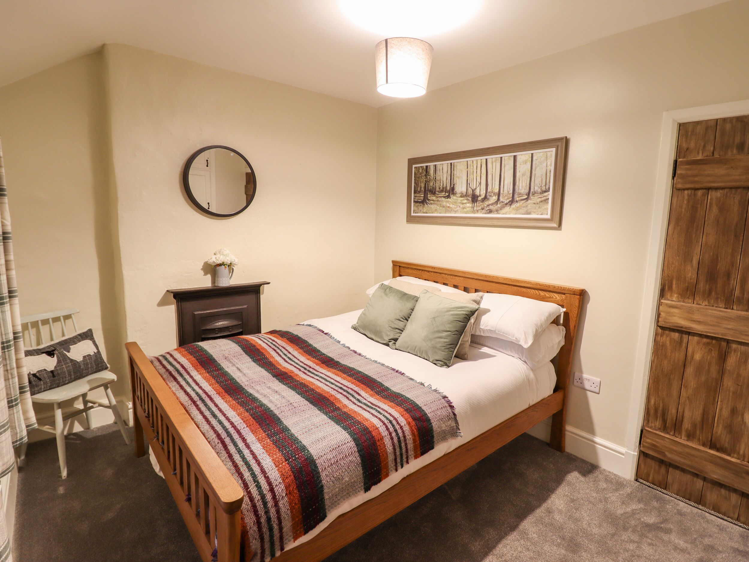 A bedroom with a wooden bed, a bedside table with a vase of flowers, a mirror, and a painting on the wall.