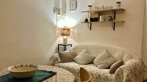 A cozy living room with a round table, a bowl, a chair, a lamp, a sofa, and shelves with tea-related items. - Gîte 2 Personnes Bruyère - 35 m² (Niozelles)