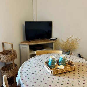 Ein Zimmer mit einem Fernseher auf einem Holzständer, einer gepunkteten Tischdecke, einem Korbkörbchen mit zwei Karaffen und einer Vase mit Trockenpflanzen.