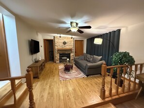 A living room with a stone fireplace, a ceiling fan, a sofa, a television, and a staircase.