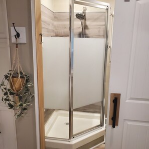 A glass-enclosed shower with a white tiled wall, a visible drain, and a macrame plant hanging on the wall. - Tiny house studio in woods, accommodates 4 people total! (Richlands)