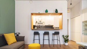 Ein modernes Wohnzimmer mit einer Couch, drei Barhocker, einem gelben Hocker und einer durch einen großen Spiegel sichtbaren Küche.