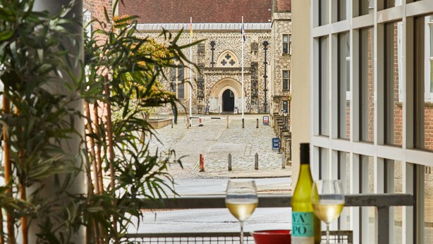 A balcony with a table, two wine glasses, and a bottle of wine. In the background, there is a building with a brick facade and a large archway. - Castle Retreat City Centre Near Train & Parking (Winchester)