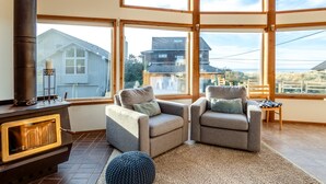 Een gezellige woonkamer met een open haard, twee fauteuils, een rond hocker en grote ramen met uitzicht op de omgeving.
