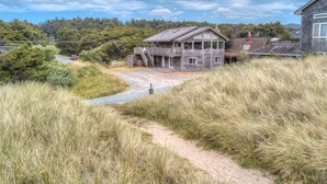Een houten huis met een veranda en een trap naar een terras, omgeven door hoog gras en bomen.