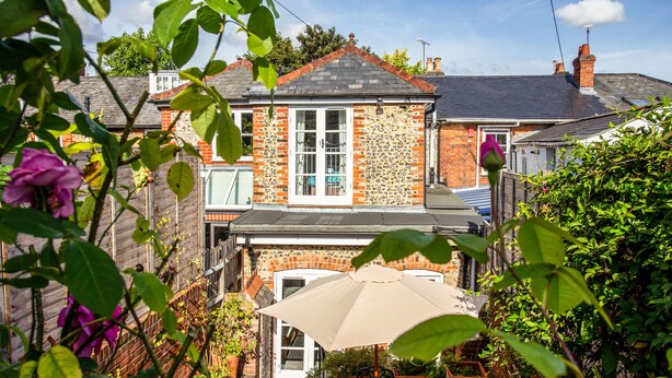 Une maison à deux étages avec une façade en briques, un toit en tuiles et un jardin devant, agrémenté de plantes en pot et d’une table de patio avec un parasol.