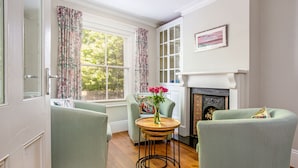 Un salon chaleureux avec une cheminée, deux fauteuils verts, une table ronde en bois ornée d’un vase de fleurs et une grande fenêtre agrémentée de rideaux à motifs floraux.
