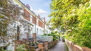Une rue étroite bordée de maisons à étages aux façades en briques et aux ornements blancs, qui mène à une entrée fermée par un portail.