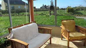 A conservatory with a wooden bench and a chair, large windows offering a view of a garden and a building in the distance. - DETACHED HOUSE 100 M2 ON A 936 M2 ENCLOSED PLOT AT THE END OF A CUL-DE-SAC (Porspoder)