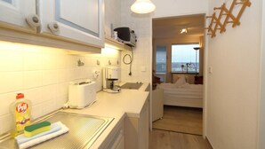 A compact kitchen with a toaster, coffee maker, and a bottle of dish soap. A bed is visible in the adjacent room, and a window offers a view of the city. - House Metropol 254, OB (Westerland)