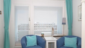 A room with two blue armchairs, a small white table, and a view through large windows with blinds.