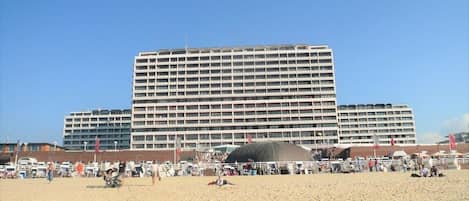 A large hotel with multiple floors and balconies, situated on a beach with a sandy foreground and a clear blue sky.