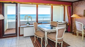 A room with a dining table, chairs, a view of the sea, and a window with curtains.
