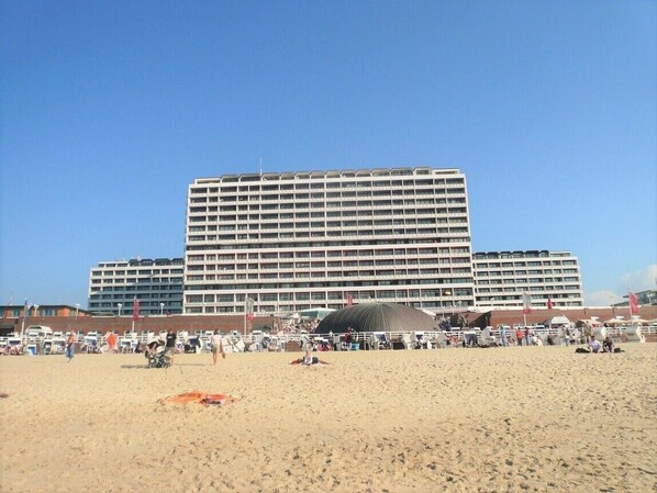 A large hotel with multiple floors and balconies, situated on a beach with a sandy foreground and a clear blue sky.