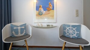 A room with two white chairs with nautical-themed cushions, a framed poster on the wall, and a small shelf with decorative items. - Haus Metropol 175 OB (Westerland)