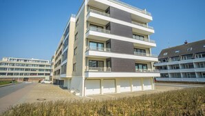 A multi-story residential building with balconies, large windows, and a parking area in front. - Haus Birgit 6-15 Steinmannstraße 15. In Zentraler und Strandnaher Lage in Westerland, Liegt Diese Gemütliche, Liebevoll Eingerichtete Ferienwohnung. Nur 100m von der Promenade/strand Sowie den Kureinrichtungen und Schwimmbad en (Westerland)