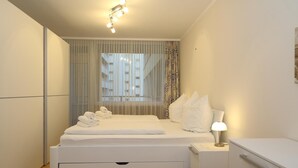 Un dormitorio moderno con cama, mesitas de noche, lámpara y vistas a edificios a través de cortinas traslúcidas.