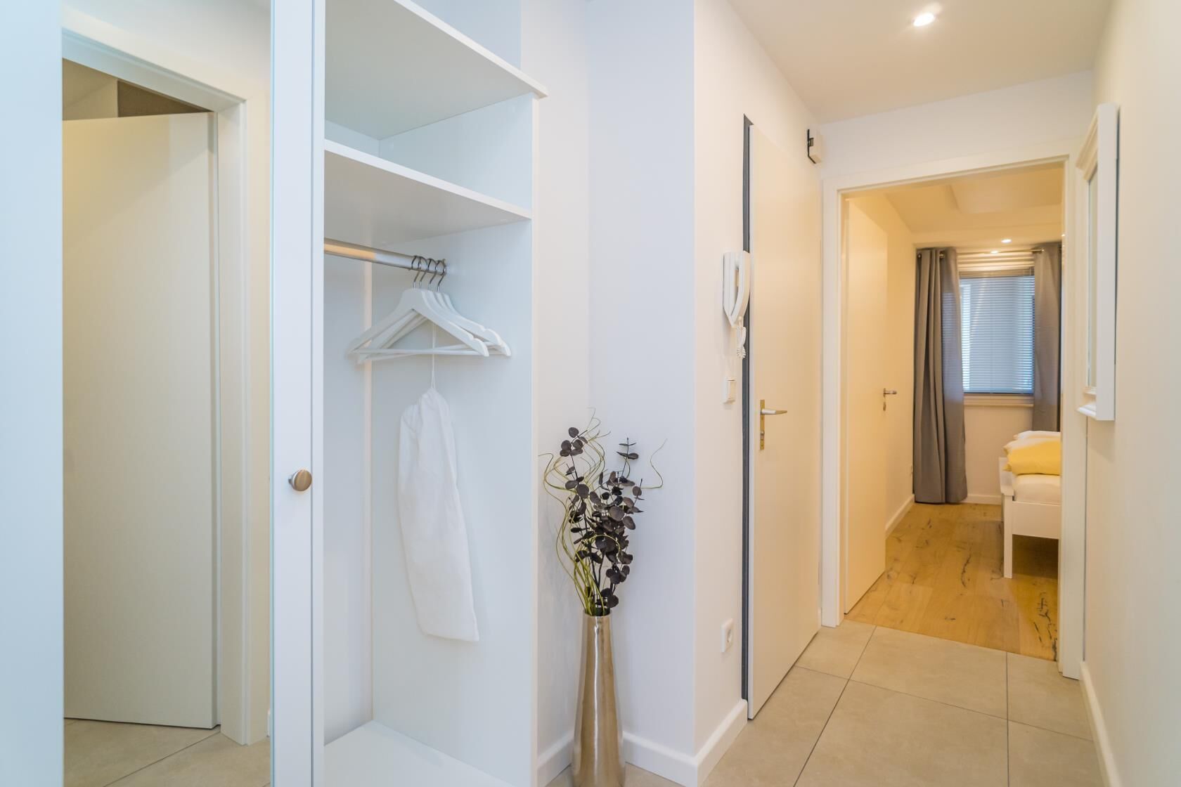 A hallway with a white wardrobe, a hanging garment, and a vase with flowers.